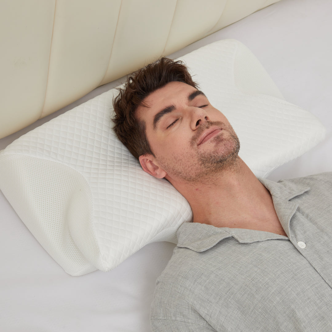 Man in a grey pajama shirt sleeping on his back, his head resting in the center of the white CozyRest pillow against a cream headboard